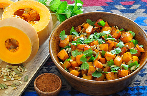 butternut squash cut in half with seeds sprinkled around next to a bowl of cooked butternut squash dish